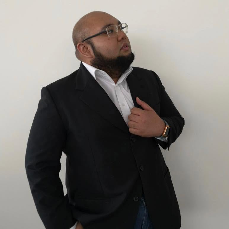 Hykarl Sufardi in glasses, a black jacket and white shirt, with one hand at his jacket.
